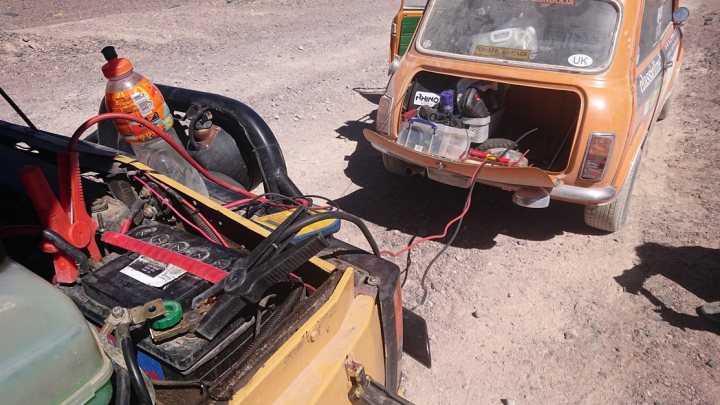 Pistonheads - The image captures a rural scene, where two older-model cars are parked on a dirt road. One car is closer to the camera and appears to be in disrepair with its hood open and several parts missing or removed. The other car, further away, seems to be intact but also old. Both vehicles show signs of use and exposure to harsh conditions. In the foreground, there's an orange truck parked, possibly suggesting some work being done on the cars. There are a few people in the scene, but they are not the primary focus. The environment suggests a desert or arid region, given the sparse vegetation and the dry, dusty ground.