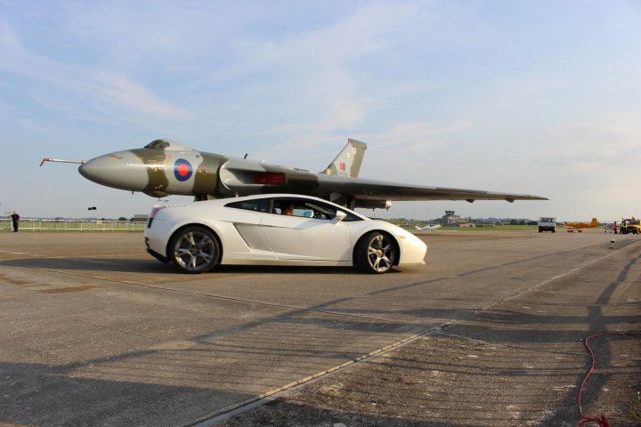 Pistonheads - The image depicts a unique pairing on a runway: a white sports car, which appears to be an Audi R8, is being towed by a fighter jet. The jet represents a modern military aircraft with its distinctive design and detailing, featuring a mix of gray and red color schemes. The runway is mostly clear with a few other vehicles visible in the background, and people can be seen in the periphery, possibly observing the event. The scene suggests this might be a demonstration or a special event involving military aircraft and high-performance civilian vehicles.