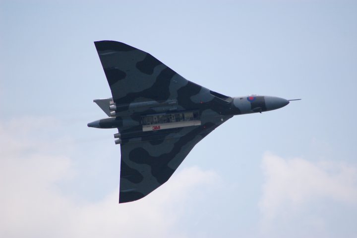 FoS Vulcan - Page 1 - Goodwood Events - PistonHeads - The image shows a military fighter jet in flight against a clear sky with some wispy clouds. The jet has a prominent sickle-shaped wing or missile, along with a second, smaller missile under its main body. Its tail section is visible with a camouflage pattern. The image focuses on the fighter jet, showcasing its sleek design and advanced technology. The clarity of the image contrasts with the complexity of the jet's design.