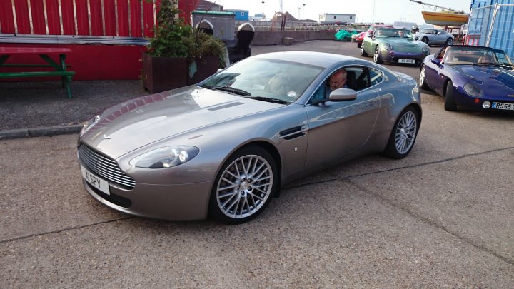 TITT 11..  - Page 23 - TVR Events & Meetings - PistonHeads - The image shows a bright silver-colored Aston Martin on a concrete surface. The car appears to be a 2-door model with distinctive sharp creases and a prominent front grille. The hood of the car is slightly raised in the center, suggesting a hardtop that might not be present in this view. There are other cars and a person in the background, but the focus and clarity of the image are specifically on the Aston Martin. The presence of the Aston Martin, usually associated with luxury and sports cars, on a concrete surface, possibly in a parking lot, suggests a casual setting rather than a race track or auto show.