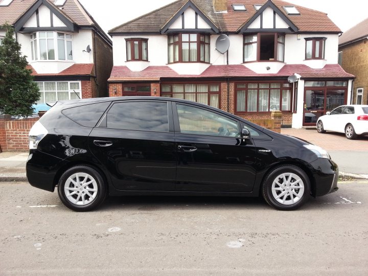 Toyota Prius Plus MPV - anyone got one? - Page 1 - Jap Chat - PistonHeads - This image showcases a black hybrid hatchback car parked on the side of a street in front of two large residential houses. The houses feature a lot of windows and terracotta-style roofing. In the background, there's a white car and some brick fences, hinting at a peaceful residential area. The black hybrid appears to be parked in a designated parking spot, and it has a set of shiny, five-spoke wheels.