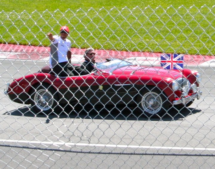 Candian GP - Page 1 - Formula 1 - PistonHeads - The image captures a moment on a racing track where two men are riding in a red vintage race car. The driver is seated in front, focused on the road, while the passenger is seated in the back, leaning slightly forward, possibly offering advice or simply enjoying the ride. The car is adorned with a British flag, displaying closed proceeding. The track is surrounded by a chain link fence, indicating a secure perimeter around the racing area.