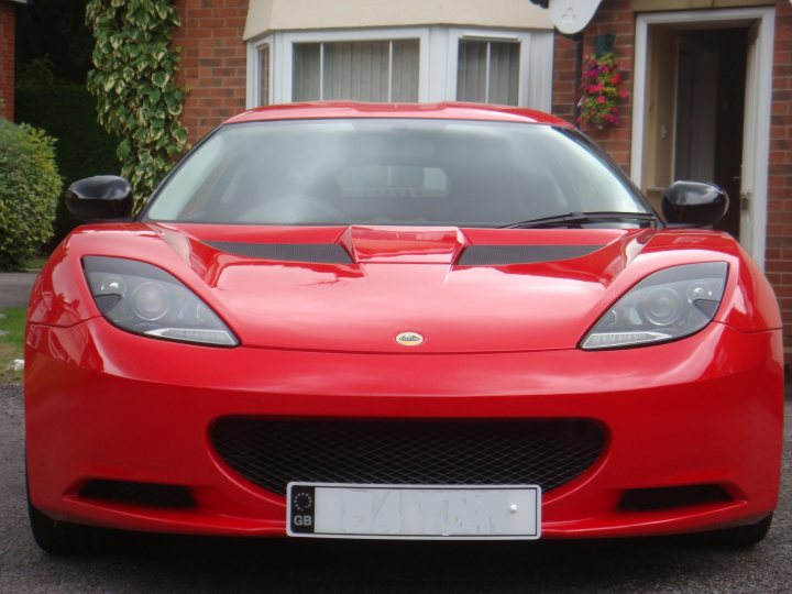 Finally some pictures!! - Page 1 - Evora - PistonHeads - The image captures a vibrant red sports car parked in front of a house. The car's sleek design and shiny exterior reflect the surrounding sunlight. The car has a black hood and black door handles, which contrast with the red body. A number plate is affixed to the front of the car, suggesting it's a registered vehicle. It's positioned on a driveway, between two long, green hedges, adding a touch of nature to the suburban setting.