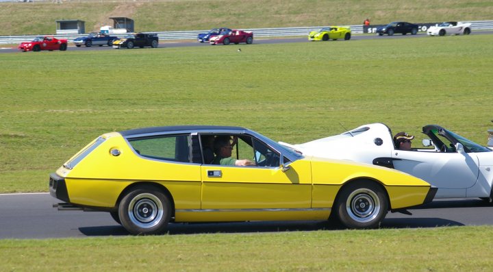 Snetterton Lotus Festival 25/26 June - Page 1 - General Lotus Stuff - PistonHeads - The image depicts an exciting scene on a race track. Two sports cars, one yellow and one white, are driving side-by-side in what appears to be an intense race. The cars are speeding around a sharp bend in the track, with the green grass of the infield visible in the background.

There's a small group of people scattered around the track, some of whom are likely to be crew members or spectators. The track itself is outlined by a series of barriers, providing a clear boundary for the vehicles.

In the distance, several other cars can be seen on the track, suggesting that this is not a run-off race but one of the more mainstream categories in motorsport. The ascending angle of the shot captures the dynamic nature of the race and the high speeds at which the cars are going.