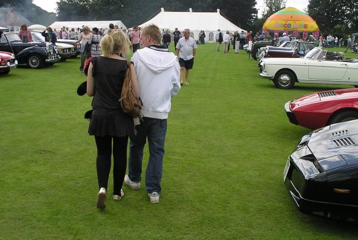 Pistonheads Sports August Brill Classic Bucks - The image captures a man and a woman walking together across a verdant field. They are passing vintage cars, suggesting they are at a car show or a similar event. The cars are turned away from the camera, but their classic designs can be discerned. The couple appears to be enjoying their time at the fair, adding to the lively atmosphere of the scene.