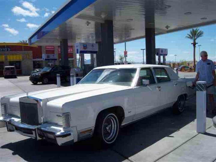 Shipping queries - Page 1 - Australia - PistonHeads - The image features a striking old-school, white muscle car with distinctive round headlights parked in front of a gas station. The car's metallic polished surface gleams under the sun, indicating it's a sunny day. Behind the car, there's a man standing next to a white gas pump, possibly preparing to pump gas. A few other cars and a truck are visible in the background, indicating a typical service station setting. The gas station has a blue and white sign, and the sky above is noticeably bright and clear, suggesting good weather.