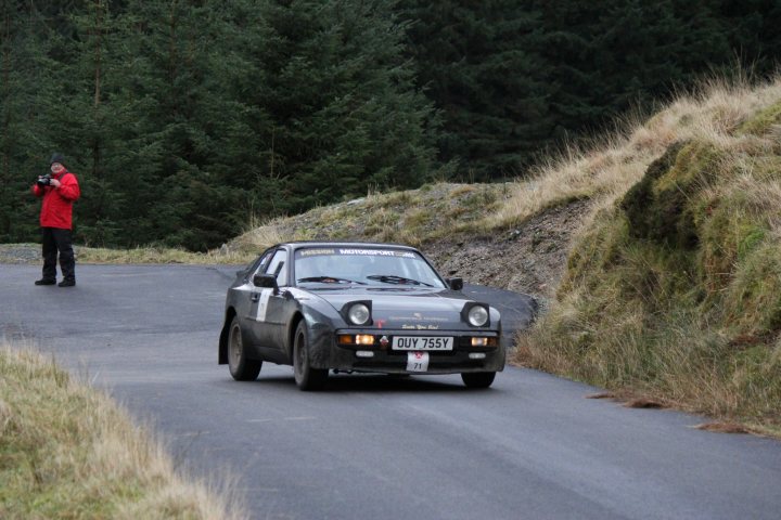 New 944 Arrival - Page 1 - Front Engined Porsches - PistonHeads - The image depicts a sharp left turn on a narrow country road, where a silver sports car is making the curve. The day is overcast with muted light, and the road appears to be made of concrete with some minor debris scattered across the surface. In the background, there's another person dressed in a red jacket standing to the left of the frame, possibly photographing the scene. The surrounding environment is lush with green trees and shrubbery, adding a natural, secluded ambiance to the scene. The sports car is weathered and carries a personal registration, suggesting it might be used frequently.