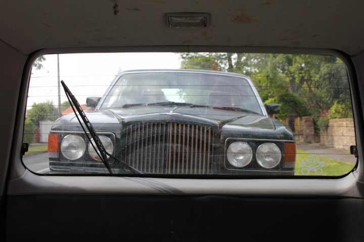 Pistonheads - This image captures a moment inside a car, where the driver's seat and dashboard are visible. The most striking feature is the front grille of a vintage Rolls Royce, which takes up a large part of the scene. The car appears to be in motion, as suggested by the blurreglass effect on the right side of the photo, which indicates a side mirror. The setting seems to be outdoors, as suggested by the presence of a tree to the left and a grass area faintly visible in the background. The overall scene is a blend of the interior of the car and a glimpse of the road ahead.