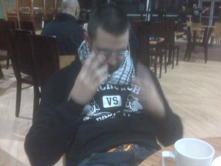 Hate Pistonheads Love - The image captures a person sitting at a table in what appears to be a restaurant. The person is wearing a black shirt with some text and graphic on it, and a bandana is being used to cover one eye. The individual is also holding a shirt with both hands, as if trying to suffocate or choke it. The surrounding area looks like a typical restaurant setting, with chairs and tables and a glimpse of another person or diners in the background. It is a candid moment, and it's unclear what the person is doing or why they're at the restaurant.