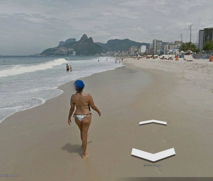 Funny things seen on google streetview UK! - Page 35 - The Lounge - PistonHeads UK - In the image, a woman is seen walking on a sandy beach next to the ocean. She's wearing a bikini and has a towel draped over her shoulders. The beach stretches out towards the horizon where buildings can be seen, indicating that the beach is located near a city. The sky above is gray, suggesting it might be an overcast day or possibly early morning or late afternoon. There are no visible texts in the image.