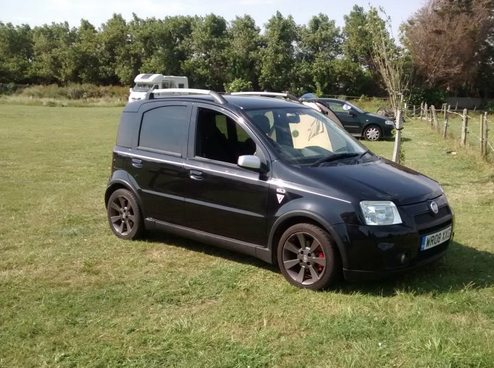 A car parked on the side of the road - Pistonheads - The image showcases a black car parked on a grassy area within what appears to be a camping site. The car is a four-door hatchback, and it features a unique symbol on the grill, which is in the shape of a cross within a circle, possibly indicative of a brand or logo. In the vicinity of the car, there are a few other vehicles, suggesting the presence of a group or several visitors. Additionally, there are structures that might be part of the camping facilities, including a small fence or partition and a tent or cabin nearby. The sky is clear and blue, suggesting fair weather conditions.