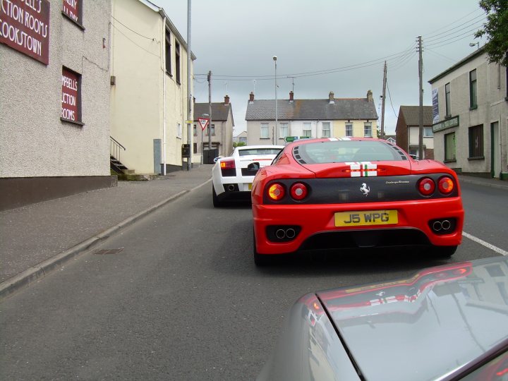 Pistonheads - The image showcases a street at dusk, with the fading light casting long shadows. Dominating the scene are two sports cars, one painted in a classic red and the other in a striking white. They are in motion, traveling from the distance towards the camera, adding a sense of dynamism to the otherwise tranquil setting. On either side of the road, building facades can be seen, their details muted by the low light. This scene beautifully captures the blend of everyday life with the occasional thrill of a high-performance automobile.