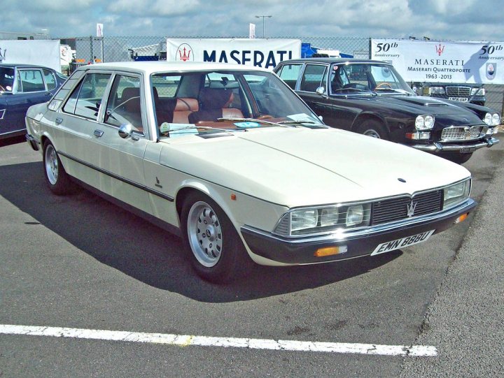 RE: De Tomaso Deauville | The Brave Pill - Page 5 - General Gassing - PistonHeads UK - The image features a vintage car, which appears to be a Mitsubishi Galant. The car is cream-colored and has a distinctive front grille with a honeycomb design. It's parked in what looks like an outdoor parking lot, surrounded by other cars, indicating it might be at a car show or event. The background suggests the location could be near a beach, given the presence of sand dunes, but it could also just be a coastal cityscape. There are no people visible in the image.