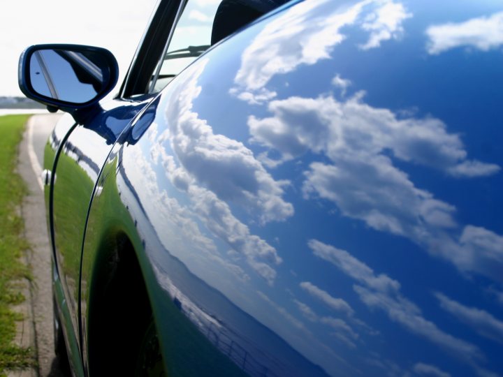 Wow! A sunny day... - Page 1 - North East - PistonHeads - The image presents a striking view of a car reflected in its shiny, blue exterior. The reflection captures a vibrant sky, the vast expanse of water, and lush, green grass. The cloud formations are also mirrored perfectly on the car's surface, mimicking the beauty of the sky above. This mirror image creates a captivating visual effect, blending the man-made object with the natural world.