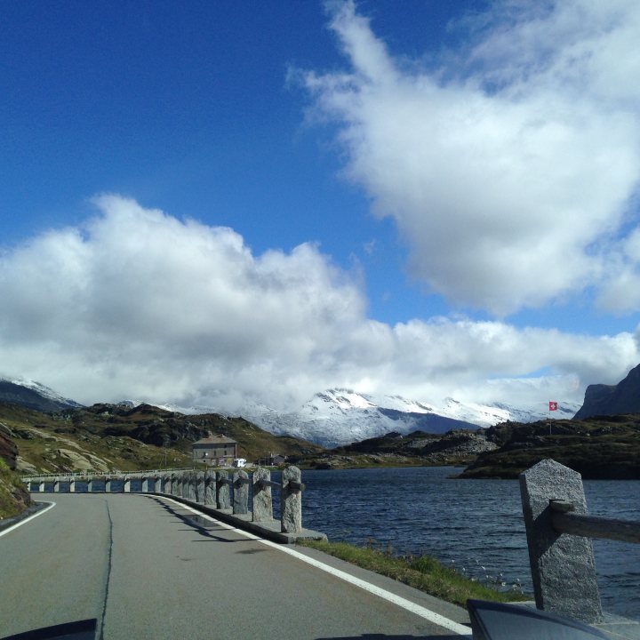 7 Countries 4 days & a banger - 'Help For Heroes' - Page 1 - South West - PistonHeads - The image shows a winding road by a lake under a clear blue sky. The road curves along the edge of the water body, and there are a few cars visible midway on the road. In the distance, a range of snow-capped mountains rises beyond the water, partially obscured by the fluffy white clouds in the sky. The overall setting appears serene and picturesque, with a prominent view that suggests it could be a touristic location.