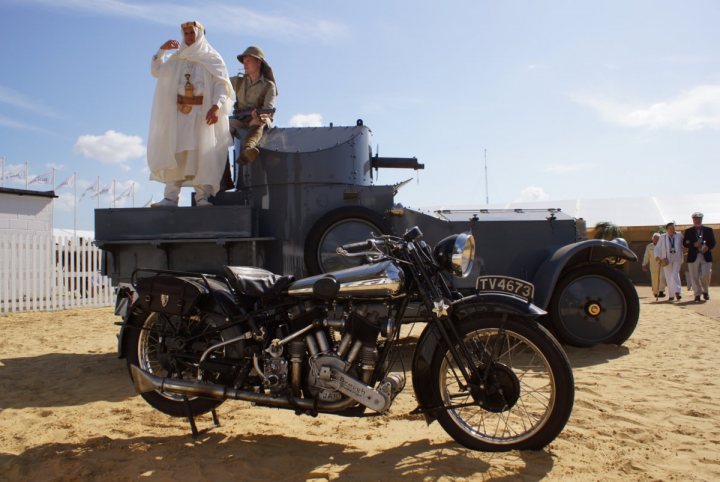 A black and white photo of a motorcycle - Pistonheads - The image depicts a scene with a man and a woman on top of a military truck parked on a sandy area. The truck is dark blue and takes up a large portion of the image. The man is wearing a white robes with a hood, while the woman is beside him. They are standing and facing the same direction, where there are a few bystanders. Additionally, a cavalry motorcycle with a prominent sidecar is parked in the foreground of the image. The bystanders and the military elements in the background suggest a ceremonial or historical setting.