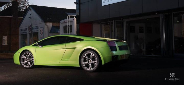 Performante, colours, numbers etc - Page 5 - Gallardo/Huracan - PistonHeads - The image shows a sleek green sports car parked at a slight angle, facing the building. The bright green sports car is positioned in front of the building, which is built with metal and concrete, giving the impression of a modern architectural style. The backdrop includes a building with advertisements, trees, and a cloudy sky, suggesting this scene could be in a city or town center. The photo has a professional and polished feel, likely intended to showcase the car. There are emblems that appear to say "Khalid it photography."