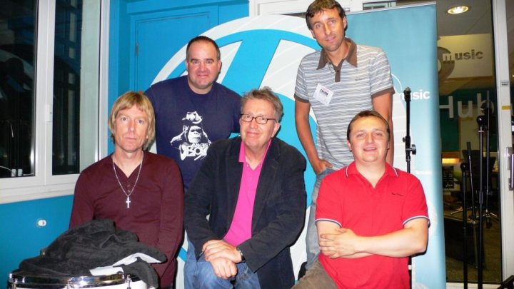 Lets Pistonheads  Gig - The image is a color photograph featuring five individuals who appear to be in a casual indoor setting. They are surrounded by music equipment and appear to be posing for the photograph. The individual on the far left is wearing a dark red long-sleeved shirt with a visible cross pendant, and to their right, there is a person wearing a black jacket over a striped shirt. He is facing the camera and his hand is on his hip. In between the two, another person is wearing a pink shirt and has his arms crossed. The individual in the center is wearing a dark suit jacket, a pink shirt, and has hands clasped in front of him. Lastly, on the far right, there is a medium-built man wearing a red shirt with short sleeves and darker hair. The background of the photo includes music-related promotional materials, soft lighting, and what seems to be a glimpse of furniture.