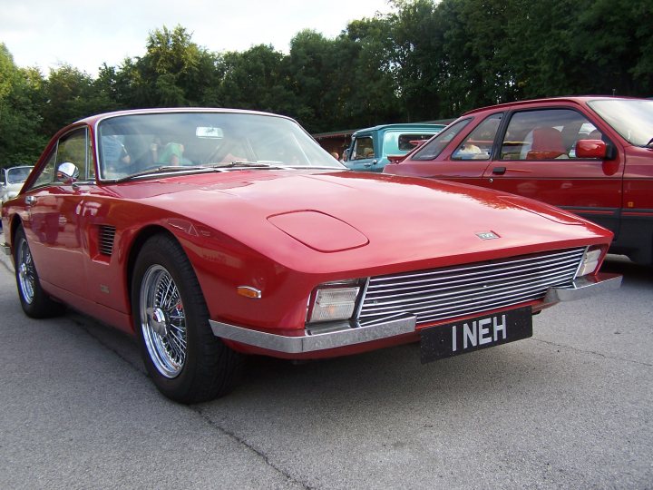 Pistonheads Trident Clipper Venturer - The image showcases a vibrant red sports car parked in a spacious outdoor parking lot. The car, which is prominently displayed and faces the viewer, appears to be in excellent condition, suggesting it is well-maintained. In the background, there are trees, adding a touch of nature to the urban setting. Beyond the trees, there are other, older cars parked in the lot, adding contrast to the image. The car is equipped with an I-NEH license plate, indicating it could be a model from the UK. The clear day provides good visibility, highlighting the details of the car and the surrounding environment.