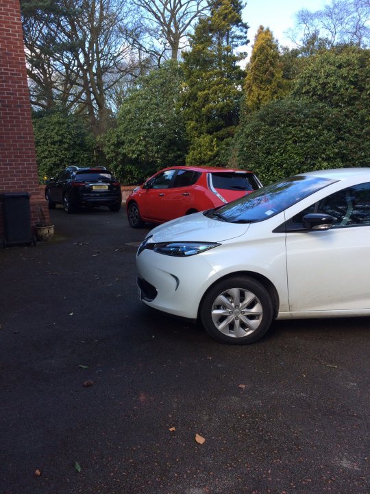 Looking at a Leaf Tekna, is this a good deal? - Page 1 - EV and Alternative Fuels - PistonHeads - In the image, there are three small cars parked in a driveway. The cars are a mix of colors, with one being a vibrant red, another a sleek black, and the third a pristine white. All three vehicles are compact, with their bodywork reflecting light, giving them a shiny appearance. The cars are positioned near a lush green tree, casting a cool, refreshing atmosphere over the scene. The driveway appears to be a residential area, bordered by a brick wall and a house visible in the background. The image radiates a sense of tranquility and orderly living.