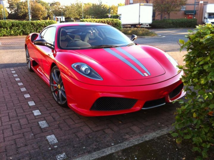 Scuderia must have options? - Page 1 - Ferrari V8 - PistonHeads - The image shows a vibrant red Lamborghini supercar driving on a road. The car's glossy finish is striking, and its design highlights its performance and prestige status. The front end is notable for its angular sculpting, sharp air intakes, and a streamlined, aerodynamic hood and front fascia. The background reveals the car to be parked in a public area with other vehicles and greenery. The setting suggests a sunny day, with the sky marked by scattered clouds.