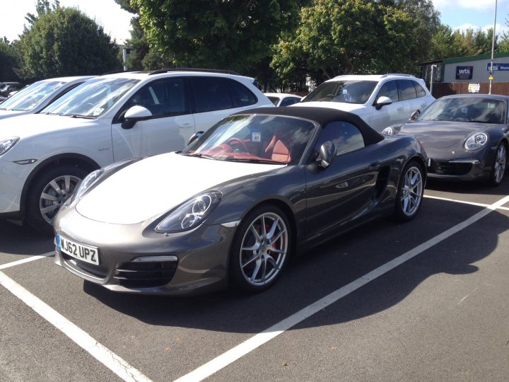 Porsche 981S Arrived! - Page 1 - Porsche General - PistonHeads - The image shows a gray convertible sports car parked in a parking lot, bordered by other vehicles, including white cars and a silver Coupe. The car has a beautiful design with a convertible top, a wide stance, and alloy wheels. The parking lot appears to be a public space, with clear markings and ample space between vehicles. The contrast between the grey car and the white cars adds a dynamic element to the scene.