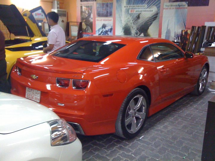 Whos Camaro Gen Pistonheads - The image captures the interior of a bustling garage or showroom, where various cars are parked. Among them, an orange Corvette sports car draws attention with its vibrant color and shiny finish. The car is positioned against the backdrop of other vehicles, showcards, and memorabilia. A man is seen standing next to the Corvette, perhaps an employee or a prospective buyer, adding a human element to the scene. The overall atmosphere suggests a place where both selling and buying dreams on wheels come true.