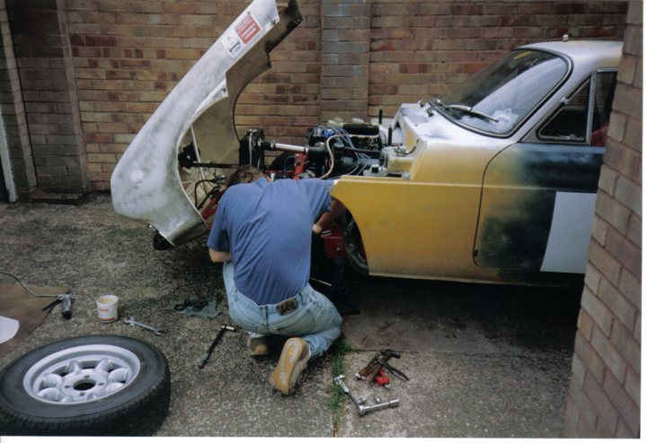 Scimitar Pistonheads Reliant - In the image, a person is working on a vintage car in a garage setting. The car is a two-toned vehicle with a distinctive hue, partially stripped, indicating some level of modification or repair work. The person is kneeling down, carefully working on the engine or possibly the underside of the car. The garage is equipped with a brick wall, adding to the vintage feel of the image. There are tools and parts scattered about, suggesting that this is a dedicated workspace for automotive mechanics.