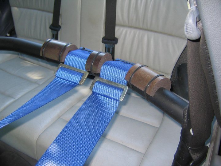 Pistonheads Harnesses Hans Device - The image shows the interior of a vehicle with a close-up view of a section of the upholstery, which appears to be leather. Notably, there are blue safety belts emerging from the backrest of the passenger seat, indicating that the car is designed to accommodate seat belts. These belts are connected to metallic attachments likely located within the seat's casing. The seat is centered on what seems to be a white floor, and there are straps attached to the backrest of the front seat adjacent to the passenger seat. The image focuses on the safety equipment, underscoring the importance of passenger safety within vehicles.