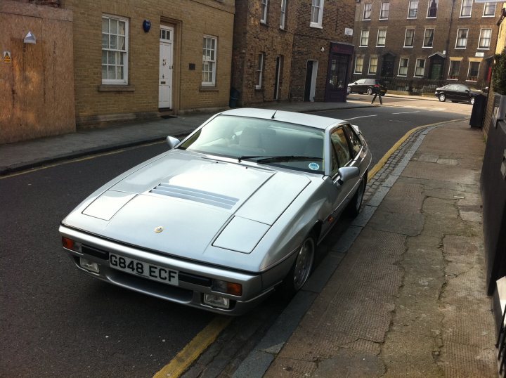 The Lotus is dead, long live the Lotus! - Page 6 - Classic Cars and Yesterday's Heroes - PistonHeads - The image showcases a silver Porsche car parked on the side of a street. The car is positioned at the edge of the sidewalk, adjacent to a curb, and seems to be facing towards the right side of the image. A yellow line is visible to the left of the car, suggesting it's a parking restriction area. The backdrop includes a brick wall and a neighboring house, hinting at a residential area. The car is the main subject in the image, with the house and the yellow line serving as contextual elements.