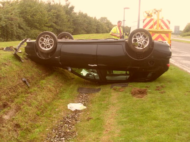 Rover and out! Cribbs crash. - Page 1 - South West - PistonHeads - The image presents a scene of a black car that has crashed and tipped over onto its roof. The car is situated on the side of a grassy embankment, and its orientation indicates a significant impact. Two individuals stand near the damaged vehicle, possibly assessing the situation. There is a yellow and white caution vehicle, most likely providing assistance and ensuring safety. The overall impression is one of wreckage and investigation.