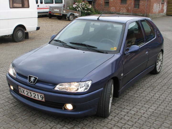 One owner FSH GTi-6 with 215K on the clock. Have I gone mad? - Page 1 - French Bred - PistonHeads - The image features a blue Peugeot 206 parked on a driveway. It appears to be a sunny day, with shadows visible on the ground. The car has a registration tag on the front plate that reads "SX 2319." In the background, there is a white van and a brick building. The vehicle seems to be an older model, judging by the design of the headlights and the style of the car body. The driveway appears to be made of stone pavers, which are somewhat worn and have different shades of grey.