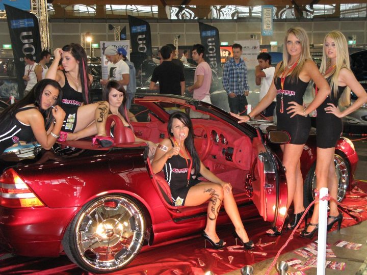 Pistonheads Sydney Autosalon - The image captures a group of women posing next to a red sports car. The car, showcased on a reflective surface, has five women inside, and two women stand outside of it. They are all dressed in black, with some sporting tattoos and one holding a drink. The atmosphere appears to be festive, with conference signs and an event setting visible in the background.