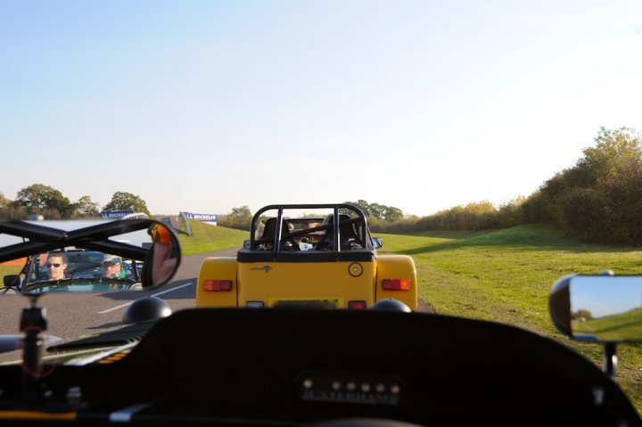 Pistonheads - The image captures an outdoor scene of a journey in progress. There is a person, possibly the driver, visible in a rear-view mirror. They are on a road, as indicated by a visible lane marker. A yellow vehicle is driving on the road towards the horizon, partially visible from the front and rear angles, suggesting it might have turned ahead of the camera's viewpoint. In the foreground, a mirror positioned on the nearest vehicle allows us to see the reflection of a number of trees along the roadside, enhancing the sense of movement and distance. The road appears to be country or rural, with its open layout and vegetation.