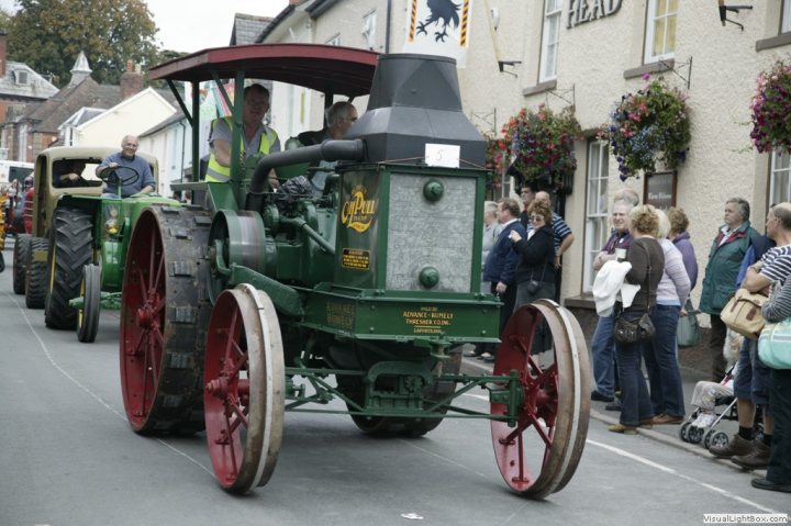 Classic tractors - Page 3 - Classic Cars and Yesterday's Heroes - PistonHeads - The image captures a vintage street scene where an old-fashioned tractor is being used to pull a wagon down the road. The tractor is green and green, with large red wheels. A man is sitting in the driver's seat, wearing a bright yellow vest, possibly operating the tractor. The wagon being pulled appears to be carrying some people, and the spectators on the side of the road are observing the tractor and wagon with interest. Some of the spectators have handbags, indicating this could be a special event or a parade. The road is lined with houses, which give a quaint village feel to the scene.