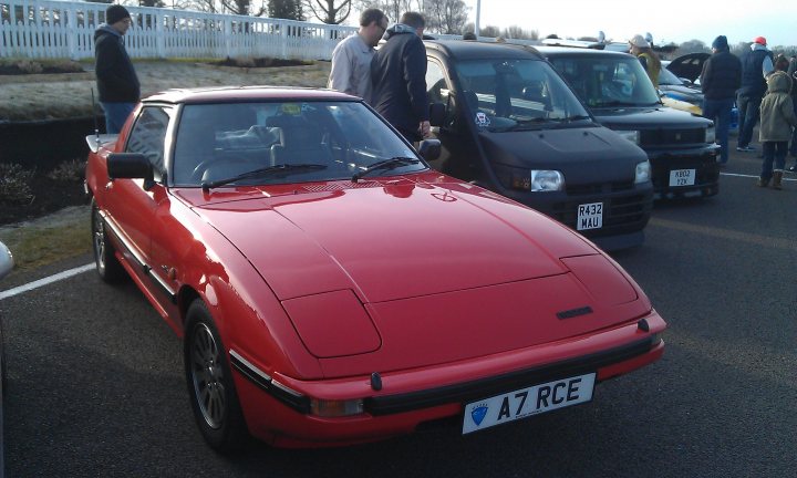 Goodwood Breakfast Club - Sushi Sunday - photos - Page 1 - Goodwood Events - PistonHeads - The image shows a parking lot where a group of people is inspecting several small red cars. The red car in the foreground is parked and has its hood up. In the background, there are more similar-looking vehicles, all in a light red color. Some of the people are standing near the cars, while others are scattered around the parking lot. There's a fence visible in the background. The sky is overcast, suggesting cloudy weather. The focus of the image is on the group and the inspection of the vehicles, indicating some sort of vehicle check or test.