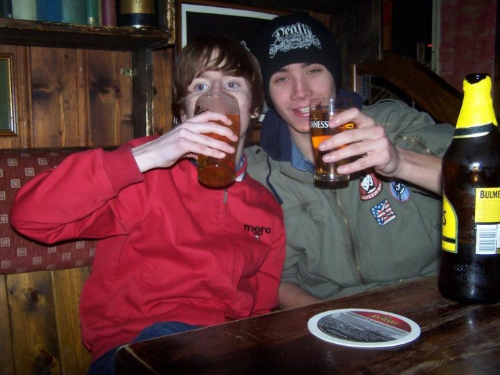 A man and a woman sitting at a table with wine glasses - The image portrays two individuals engaged in a convivial moment, sharing a drink at a bar or similar setting. One is in a red sweatshirt and the other in a black and yellow ski cap, suggesting different weather expectations for their outings. They each hold a glass filled with a golden beverage, perhaps beer, with the setting illuminated by the soft glow of the bar behind them. The camaraderie they share is underscored by their identical poses and the relaxed ambiance of their surroundings.