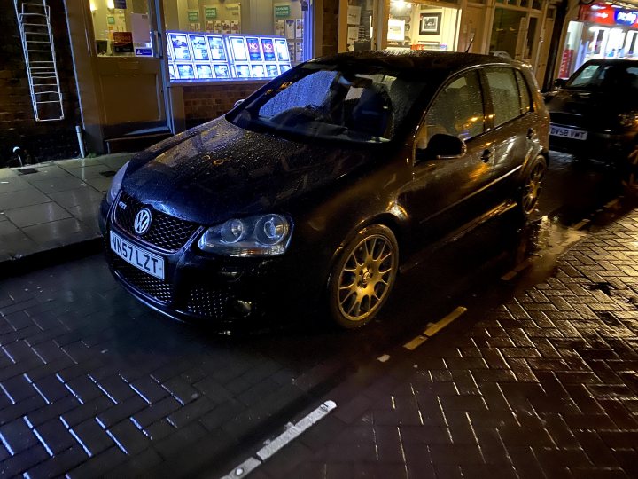 Stage 2+ Shopping Trolley - GTI Edition 30 Content - Page 1 - Readers' Cars - PistonHeads - The image captures a city scene at night. A black car is parked by the side of a street, under the protective cover of a parking lot. The car features white rims and a distinctive yellow stripe. The scene is illuminated by the lights of nearby buildings and cars, adding to the urban atmosphere. Despite the darkness, the reflections on the wet surface suggest that it has recently rained.