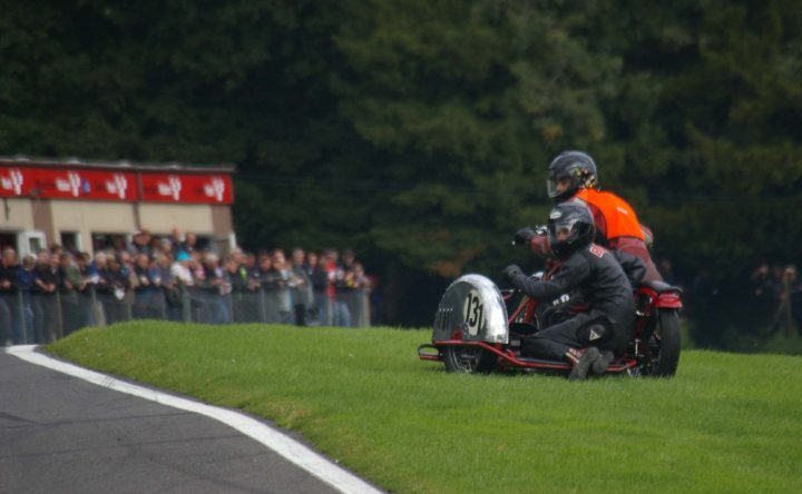 Vintage Motorcycle Club race pic's - Cadwell - pic heavy! - Page 1 - Biker Banter - PistonHeads - The image captures a dynamic scene on a curved race track. Two motorcyclists, both dressed in black and orange attire, lean low on their bikes, expertly navigating the turn. The rider in the lead closes their throats, indicating high speed, while the second rider follows closely behind, their wheels angled into the curve. In the background, spectators line the edge of the track, their focus riveted on the racing action. Above them, trees stretch towards the sky, providing a stark contrast to the industrial cranes dotting the landscape. The entire scene is a thrilling snapshot of motorcycle racing on a sunny day.