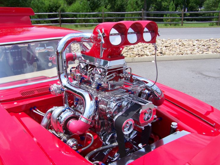 Pictures of decently Modified cars [Vol. 2] - Page 75 - General Gassing - PistonHeads - The image shows a red, classic muscle car with the hood open, allowing a clear view of its silver, stainless-steel engine. The engine features an intake manifold, headers, and the shiny, chrome components typical of such a vehicle's enhancement. The car is sitting on a concrete surface, possibly a driveway or a parking lot, and there is a grassy area with trees in the background. The faux side pipes, a common upgrade on these types of cars, are also visible on the side. No text is present in the image. The vehicle's design and modifications suggest that it may be used for racing or custom car events.