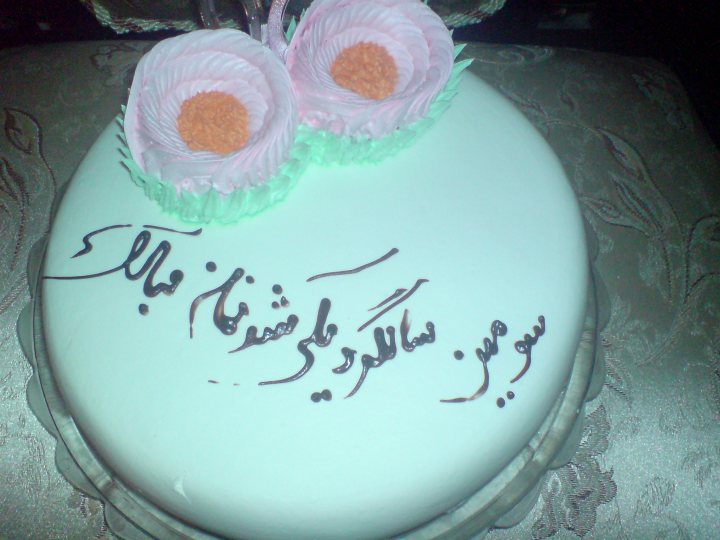 In the image, there's a beautifully decorated round cake placed on a table. The cake's frosting is white and adorned with several pink flowers, adding a touch of elegance. The most striking feature is the congratulatory message in Arabic script, cut out of the frosting, suggesting that it's a celebratory occasion. The precise placement of the flowers and the careful crafting of the text give the cake a personal and thoughtful touch.