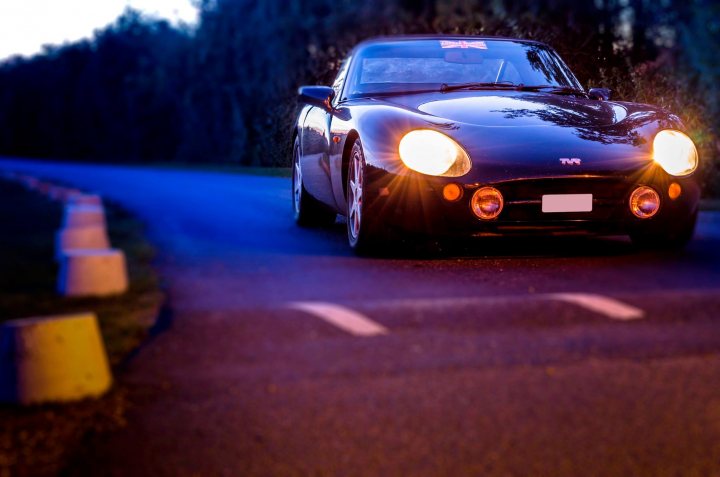 *** Essex PH Meet Thursday 15th October 19:00 *** - Page 1 - Kent & Essex - PistonHeads - This image features a black sports car positioned on the right lane of a road, as indicated by the lane marker's arrows. The car is in motion, with its headlights on, suggesting it's either early morning or late at night. The surrounding environment is dark and cloudy, and there is no text visible in the image. The setting appears to be that of a secluded countryside road.