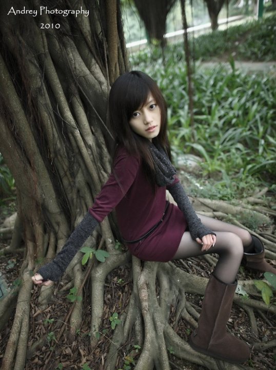 This image captures a tranquil scene in a forest. The central figure is a young woman with deep brown hair. She is dressed in a maroon dress and matching brown boots. She sits comfortably with her hands resting on a tree trunk that appears to be hollowed out by time. The background is filled with lush green foliage, suggesting the season is likely spring or summer. The woman's peaceful demeanor and the surrounding natural beauty evoke a sense of serenity and connection with nature.