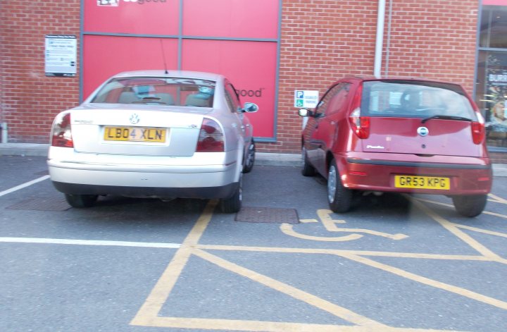 The BAD PARKING thread [vol3] - Page 324 - General Gassing - PistonHeads - The image shows a designated parking area for disabled drivers. Two vehicles are parked in these spaces, a gray car and a red SUV, both with visible license plates. The disabled parking spaces are marked by a yellow outline, and there are no passers, suggesting these spaces are being used for their intended purpose. The time on the vehicle's registration will soon expire, visible on the right window of the gray car. Behind the vehicles, there is a brick wall, likely part of the building adjacent to the parking area.