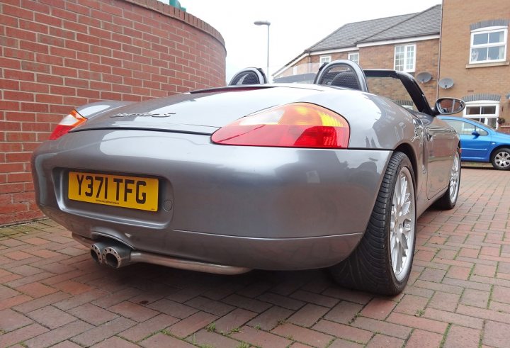 Elderly Boxster S - Page 1 - Readers' Cars - PistonHeads - The image shows a dark-colored sports car parked on a red brick pavement. The car has an elegant and sleek design, featuring a convertible top that is currently raised, providing protection from the elements. The number plate prominently displays "Y37 IGF," suggesting it's registered in Great Britain. The scene is framed by a brick wall on the left, with a clear blue sky forming a beautiful backdrop to the street where this sports car is parked.