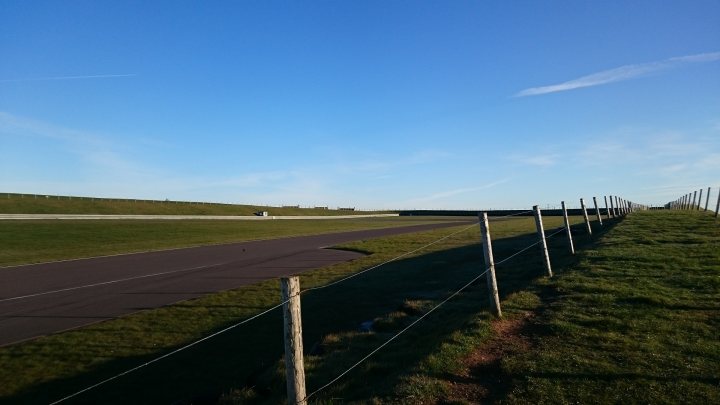 the bb trackday thread.   - Page 30 - Biker Banter - PistonHeads - This is a photograph of an outdoor scene, set in daylight. A fence with multiple vertical supports runs along the right side of the image, encapsulating a large grassy field that stretches into the distance. The field is bordered by a paved racing track or open roadway curving to the left on the opposite side. Above, the clear blue sky suggests a sunny day. The style of the photograph is realistic with no visible signs of artistic alteration.