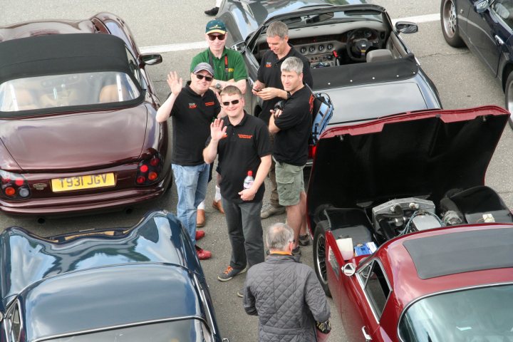 A group of people standing around a truck - Pistonheads - The image captures a gathering of men in a parking lot filled with various cars. They are dressed in black shirts with logos, which suggests a group or possibly a car gathering or event. One man is actively engaging with the others, creating a sense of casual interaction among the group. The vehicles in the lot vary in model and color, indicating a public or non-organized gathering. The men are standing close to the cars, possibly discussing something related to the vehicles or sharing their enthusiasm for cars in general.