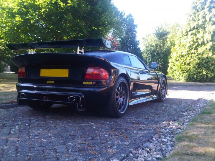 Noble Pistonheads Gto - The image shows a sleek black sports car with the top down, parked on a driveway. It has a distinctive rear spoiler and yellow license plate. The concrete driveway is lined with grass and leads to a well-manicured lawn with a backdrop of lush green trees, suggesting a residential area. The car's position on the gravel and its location in front of a tree highlight its presence and add to its aesthetic appeal.