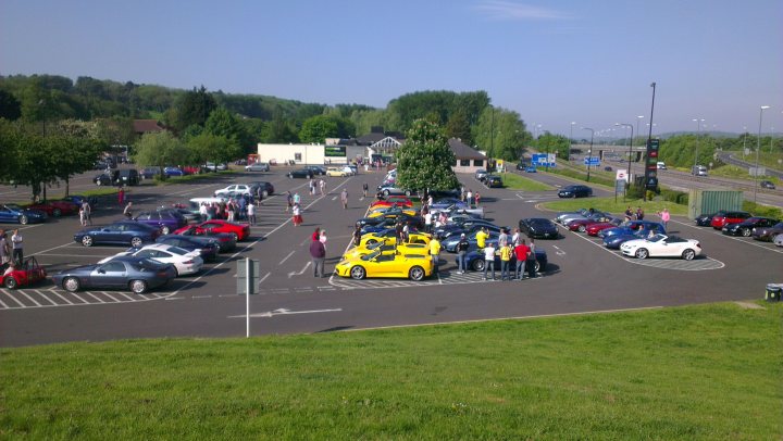 Gordano this morning en route to cwc - Page 1 - South West - PistonHeads - The image shows a lively parking lot bustling with activity. A yellow car stands as a central attraction, surrounded by several other vehicles including cars and trucks. A number of people are scattered throughout the scene, some standing near their vehicles, while others seem to be walking around the lot. The parking lot is situated in an urban setting, adjacent to a highway and surrounded by trees. Overall, the scene conveys a sense of movement and community.