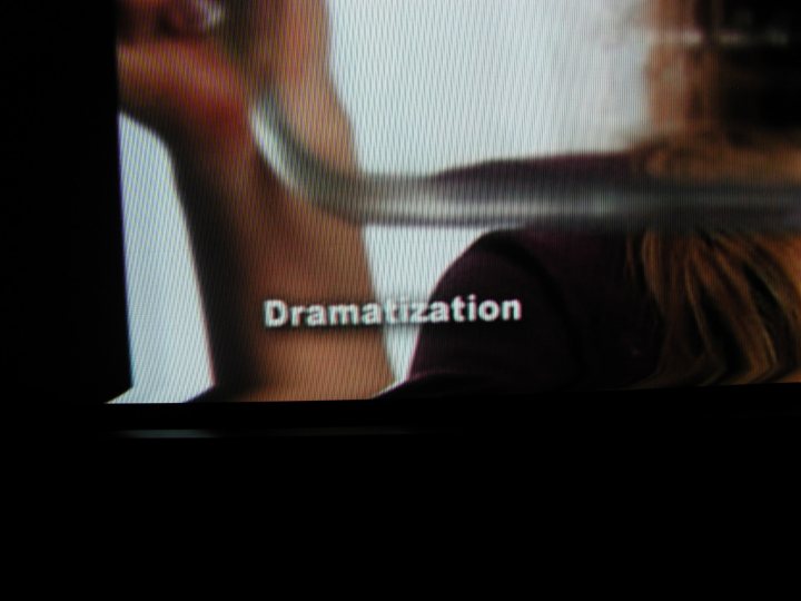 The image displays a close-up detail on a television screen. It shows a human arm in profile, held up to the screen. The foreground is somewhat blurry, but the word "Drama" is visible, suggesting the content being played is a performance or television show. The style of the image is documentary, capturing a candid moment from an ongoing event. The background is unclear due to the motion blur and the focus being on the text and the person's arm.